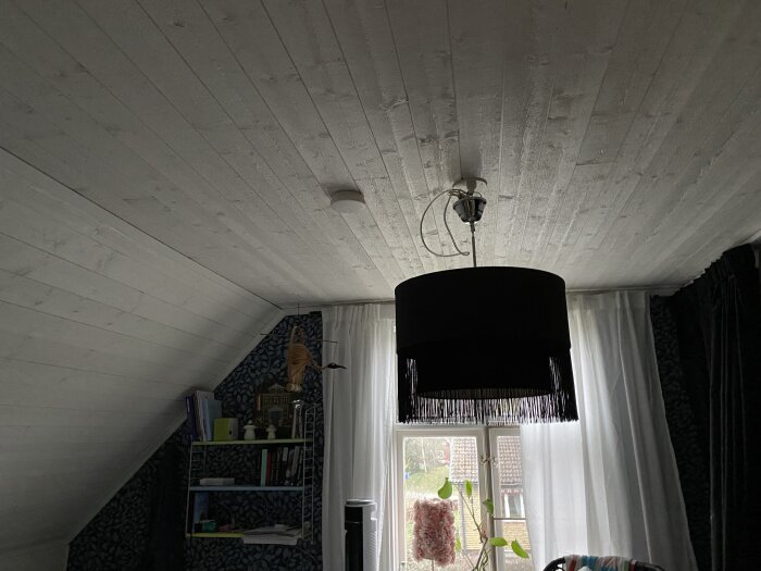 Brandvarnare i taket i ett rum med snedtak, svart taklampa och fönster med vita gardiner.