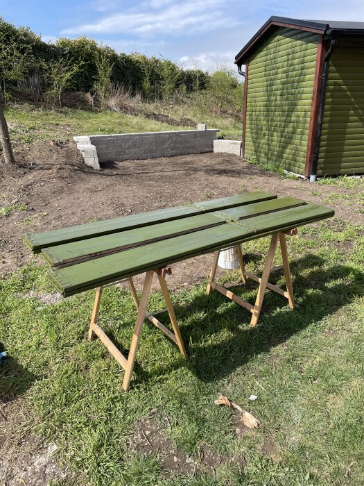 Gröna plankor torkar på bockar framför ett grönt skjul i en trädgård med nylagd mur och gräsmatta.