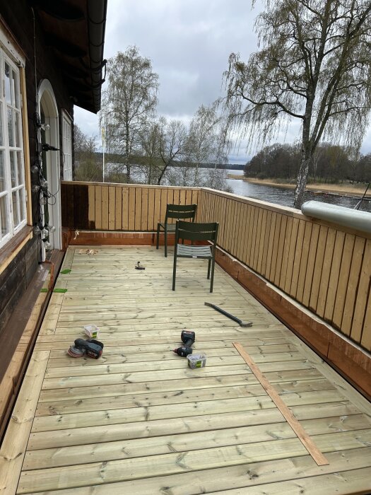Nybyggd balkong med trätrall, verktyg, och två stolar. Utsikt mot träd och sjö. Klar för sommarens utevistelse.