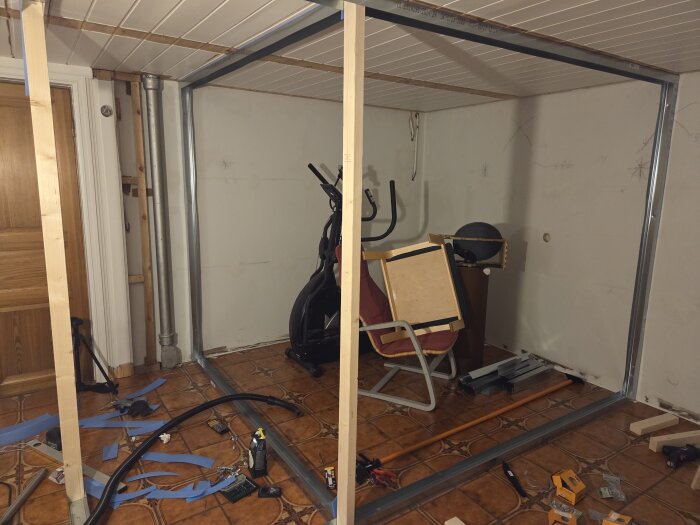 Rum under konstruktion med stålskenor monterade på golv och tak, träreglar, verktyg och en hopfälld stol.