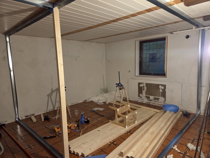 Byggprojekt i rum med stålskenor och träreglar på golv. Verktyg och byggmaterial utspridda. Fönster med enkelt glas och hantverkarbock synliga.