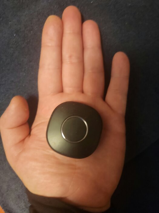 En hand håller en liten svart, rund fyrkantig enhet med en cirkel i mitten på en mörk bakgrund.