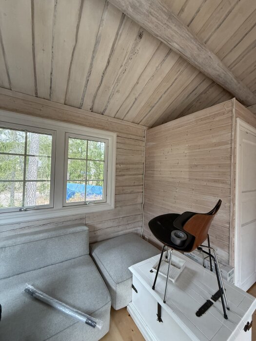 Invändigt rum i timmerhus med ljusa väggar, fönster med utsikt och hörnsoffa; modern stol på vit kista.