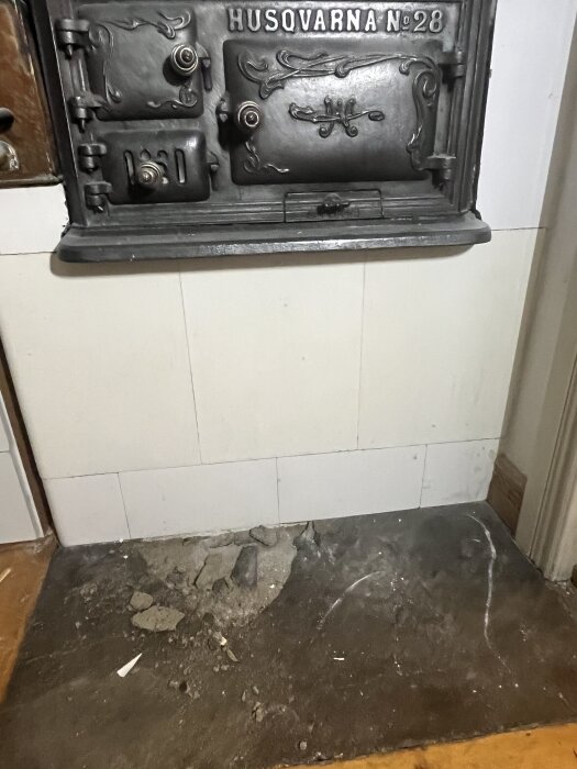 Svart eldstadsplan för vedspis med vittrande betongyta framför Husqvarna spis, grått underlag synligt på vissa ställen.