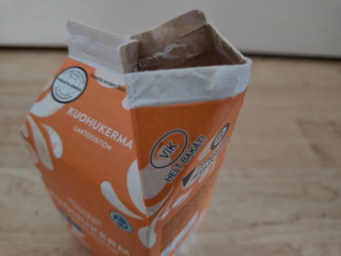 Orange mjölkförpackning från Finland märkt "Kuohukerma Laktoositon" med öppnad flärp, på trägolv.