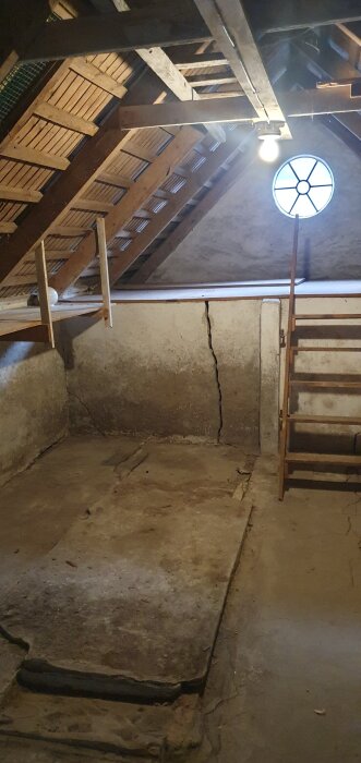 Vindsutrymme med betonggolv och sprickor, träbjälkar i taket, runda fönster, trappa och synlig vägg med sprickor i gammalt hus i Skåne.