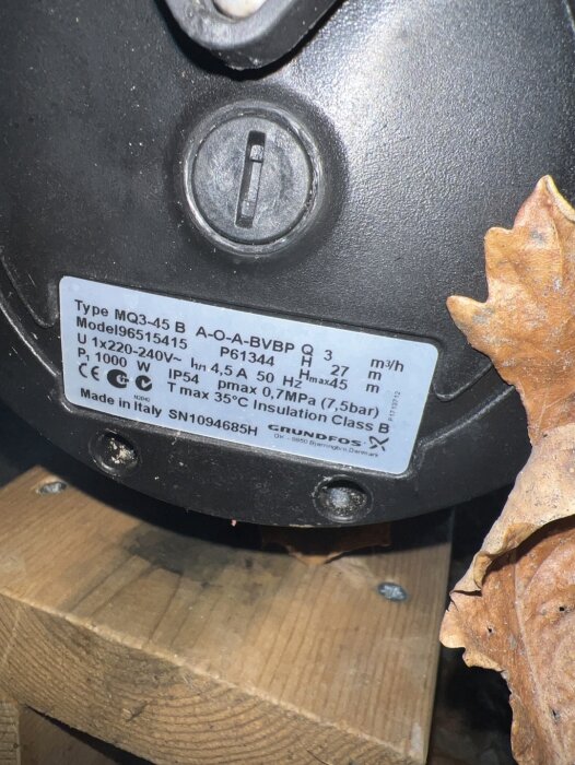 Grundfos pump MQ3-45 B med specifikationer på etiketten, placerad på en träyta bredvid höstlöv.