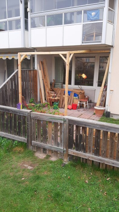 Pergola med två pelare och oavslutat tak, virke och verktyg ligger runt träddäck inhägnat med staket, lampan lyser inomhus.