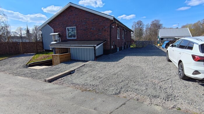 Gruslagd uppfart och gård vid ett tegelhus, med målad träpanel och parkerad vit bil under blå himmel.