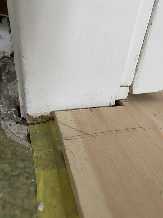 Golv med furulamellbräda nära dörröppning, med glipa mot dörrkarm. Mätlinjer syns på brädan. Vitt dörrkarm trä, murad vägg och golvsockel.