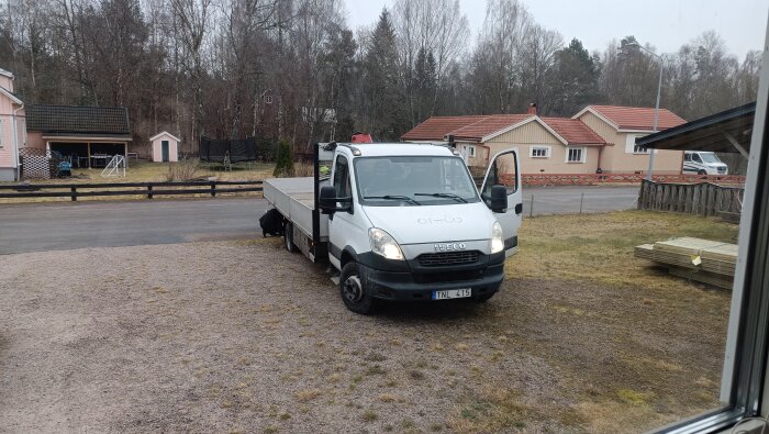 Vit lastbil med öppen dörr på grusuppfart framför hus, omgiven av träd och ett hägn, med leverans av trä bredvid.