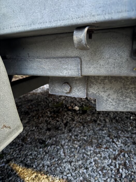 Bult fastsatt i ett grått metallbeslag under ett släp, omgiven av grusmark. Bulten verkar vara problematisk för tippfunktionen.