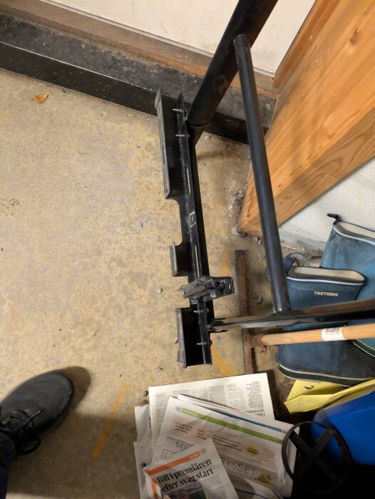 Svart metallkonstruktion vid vägg med steg och urtag, omgiven av tidningar och en stövel i förvaringsutrymme eller verkstad.