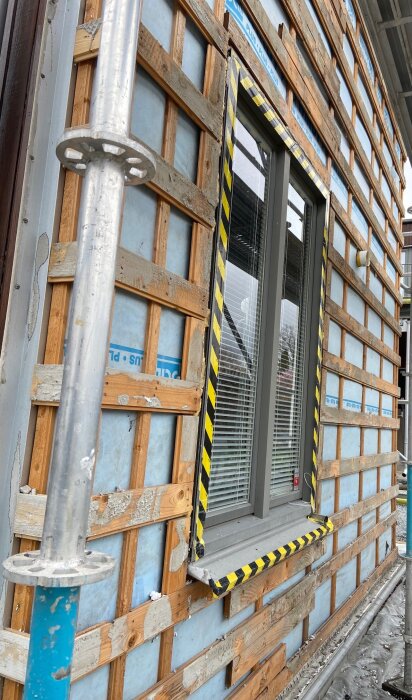 Fasadrenovering av hus med borttagna putsskivor och exponerad trästomme. Ett fönster med skyddande tejp syns. Byggställning intill.