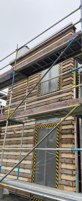 Byggställning framför ett hus under renovering med borttagen puts och cellplast. Träpaneler och plastöverdrag syns på väggen.