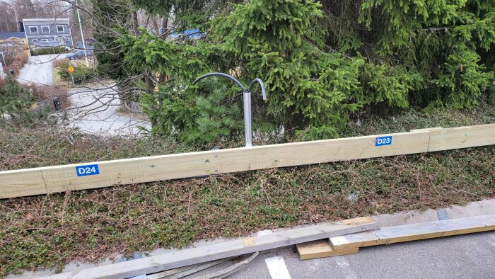 Trästolpe med etiketter "D24" och "D23", omgiven av buskar och granar, synlig på en trottoar nära en väg.
