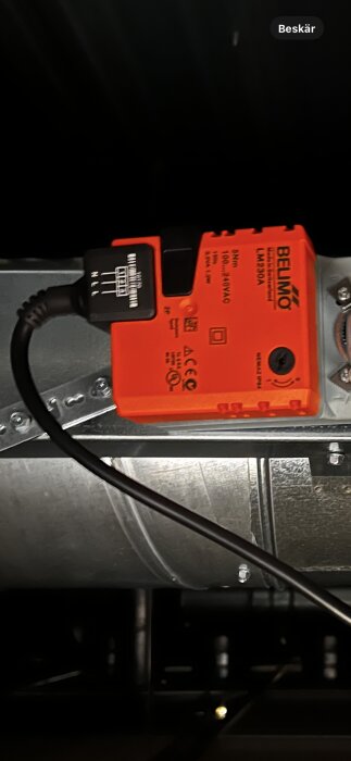 Orange Belimo spjällmotor kopplad till en kanal, med svart elanslutning, synlig kabel och mekaniska detaljer.