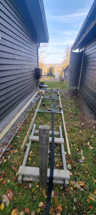 Träram med båtrullar och vinsch mellan två hus, byggmaterial och verktyg synliga i gräset.