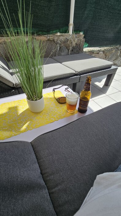 Ölglas och San Miguel-flaska på utebord med gul duk och solglasögon, växt i vit kruka, utomhussoffa med grå dynor.