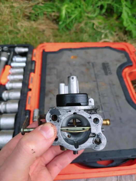Person håller en förgasare upp och ned, med synlig nålventil och choken aktiverad. Verktygslåda i bakgrunden.