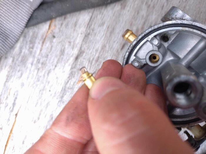 Närbild av en förgasare där en hand håller flottörnålen över förgasarhuset, med bränsleinloppet tydligt synligt.