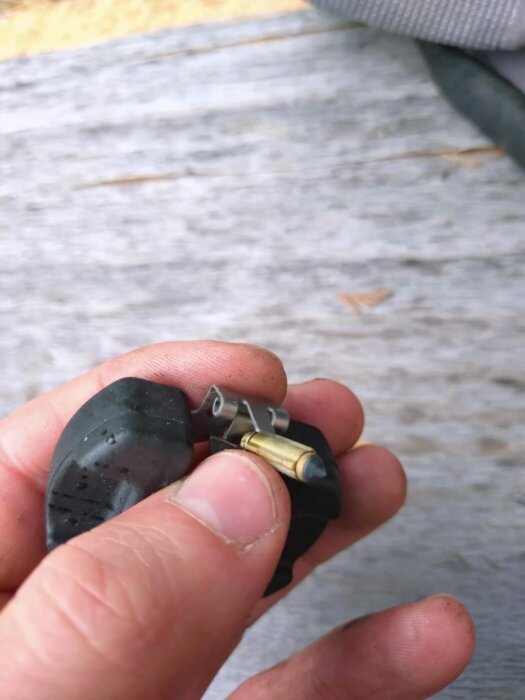 En hand håller i en bränsleflottörnål med fjäder från en demonterad förgasare på en träbakgrund.