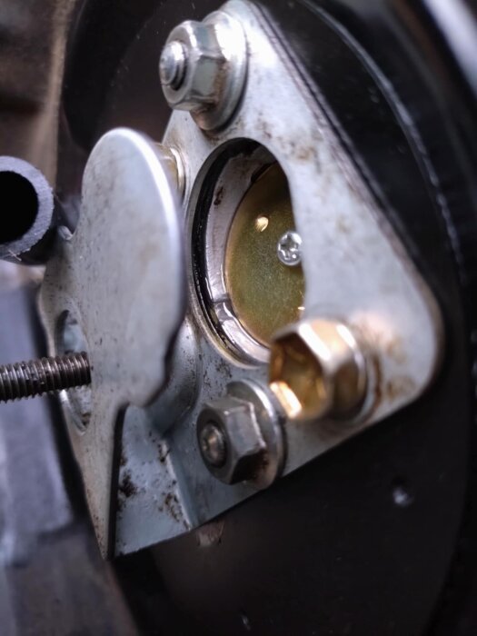 Flottörnål monterad i en förgasare, omgiven av metallskruvar och muttrar, med flottören i bakgrunden.