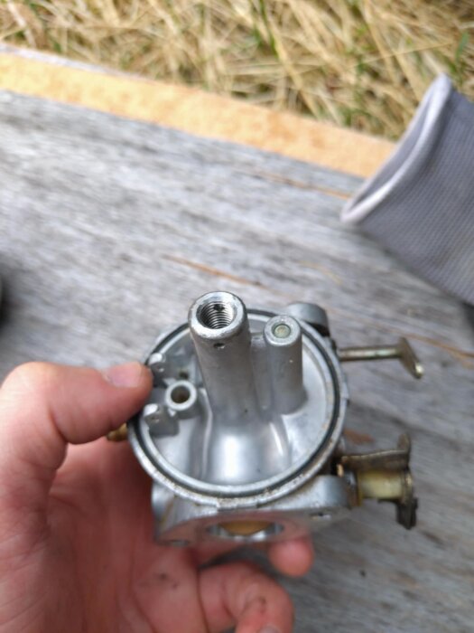 En person håller i en bildel, förmodligen en förgasare, med flera öppningar och metallkomponenter synliga, troligen relaterad till flytnålens funktion.