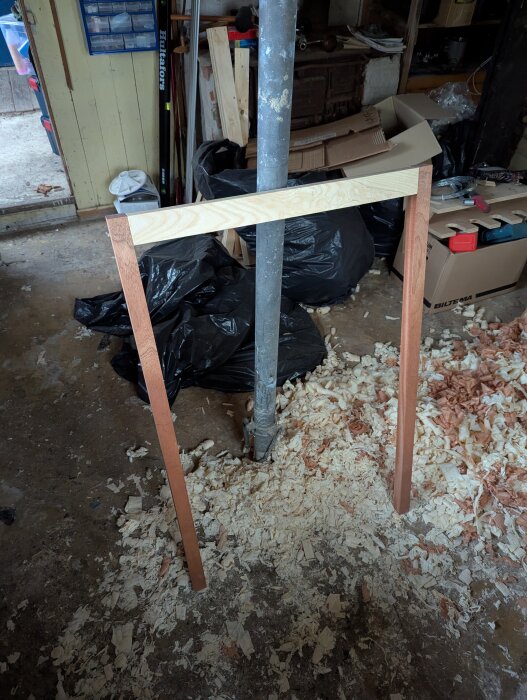 Träram med tapphål på marken i en verkstad, omgiven av träspån och verktyg. Svarta sopsäckar och kartonger i bakgrunden.