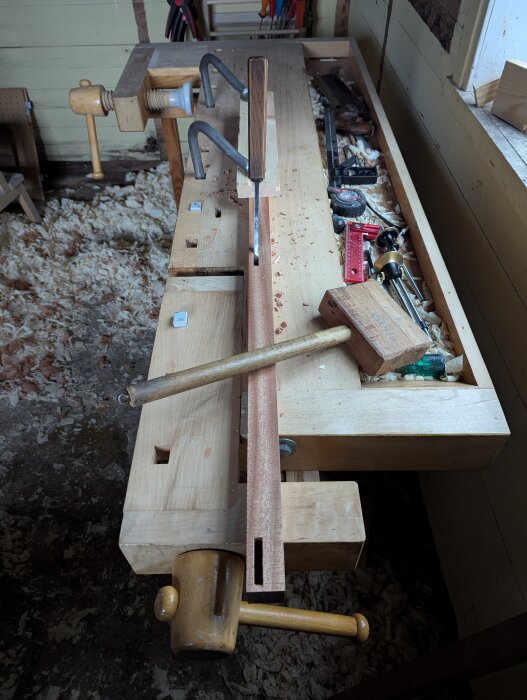 Arbetsbänk med träverktyg, inkusive hammare och markeringar för tapphål i trä. Spån på golvet i en rustik verkstadsmiljö.