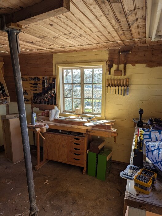 Ett rustikt skjul med arbetsbänk, verktyg på vägg och fönster som släpper in naturligt ljus, omgivna av träpaneler.