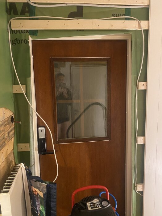 Dörr i trä monterad mot en vägg med ångbromsduk. Elektriska installationer synliga runt dörrkarmen med en grön plastfilm på väggen.