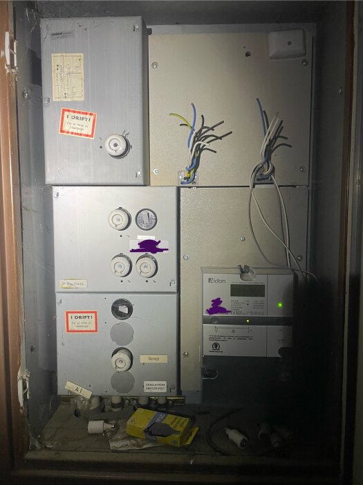 Elskåp med mätare och oskyddade kablar, märkta paneler och förvaringslådor för säkringar synliga.