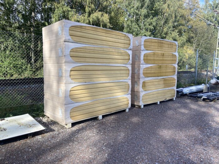 Staplade isoleringsskivor på en grusplan vid ett stängsel, omgivna av träd.