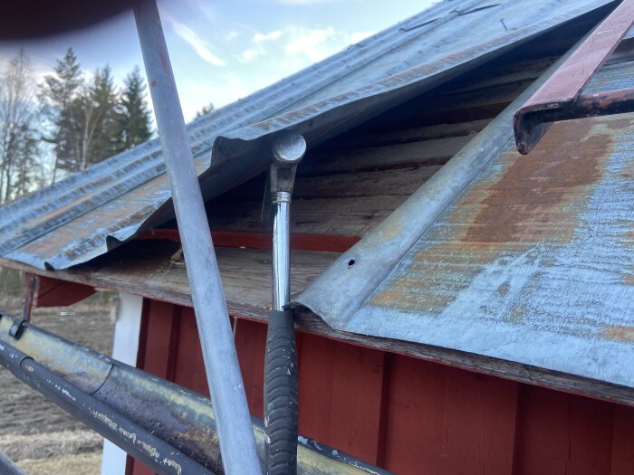 Bild av ett gammalt pannplåtstak delvis avlägsnat för reparationsarbete, med ett rödbrunt trähus och skog i bakgrunden.