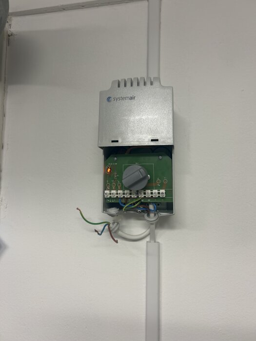 Väggmonterad Systemair-transformator med öppen front och synliga kopplingskablar samt en kontrollknapp på kretskortet.