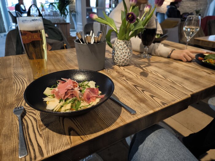 Svart tallrik med pennepasta, sallad och Serranoskinka bredvid ett glas öl på en träbord i en restaurang.