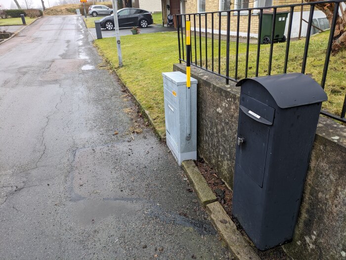 Svart postlåda och elskåp på trottoaren vid sidan av en asfalterad väg nära ett hus med gräsmatta och staket.