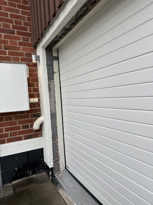 Vit garageport bredvid tegelvägg och stuprör, visar öppningens kant och blodfärgade paneler ovan.