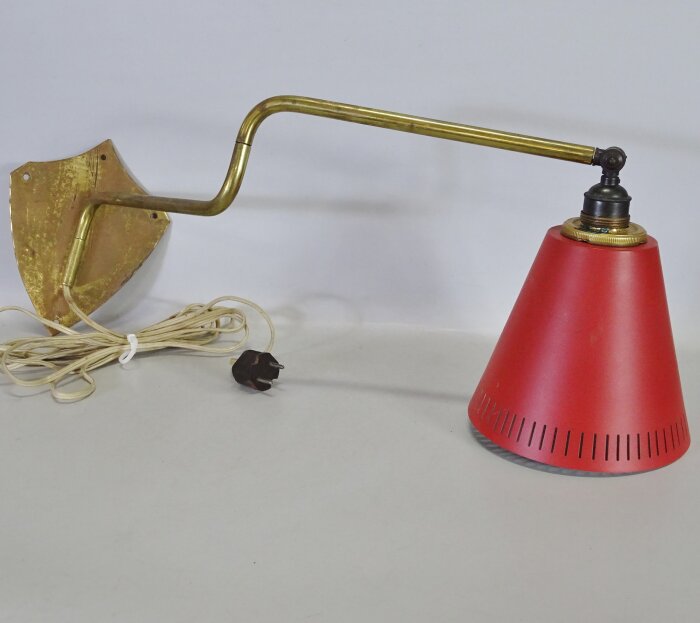 Väggmonterad lampa med röd konformad lampskärm och metallarm. Textilkabel och trepolig stickkontakt.