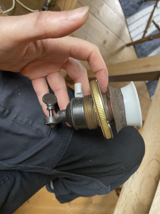 En hand håller en gammal lampanslutning av mestadels metall med porslinssockel och ring. Lampan ska förnyas, behålla originaldelar, och byta till SKX-kabel.