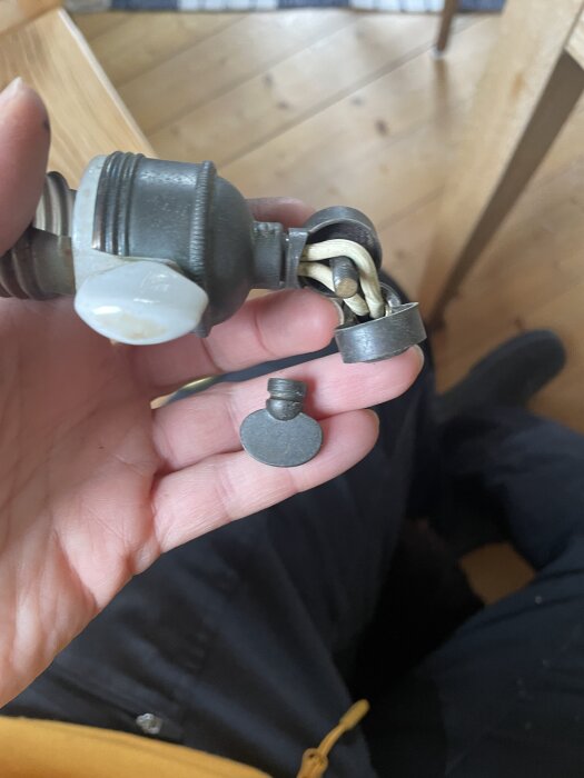 En hand håller en gammal lampa med metallkomponenter och sladdar, inklusive en porslinssockel och porslinsring, som ska få ny kabeldragning.