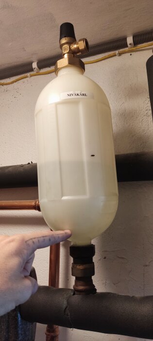 Finger pekar på ett vit plastnivåkärl i ett värmesystem, omgiven av rör och ventiler, installerat 2015.