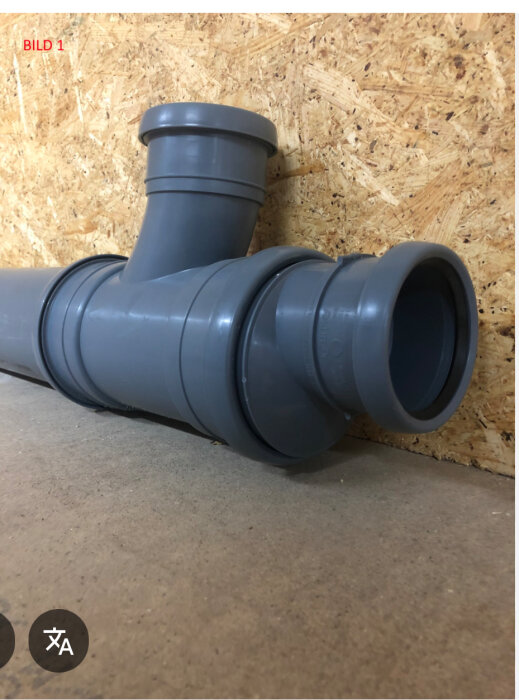 Grå plastavloppsrör med förminskning uppåt, monterat mot en vägg av träskivor.