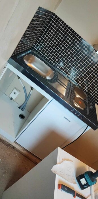 Trinettkök med rostfri diskbänk, spishäll, kylskåp under och svartvitt kakel på väggen. Verktyg och papperslapp på hörnbänk.