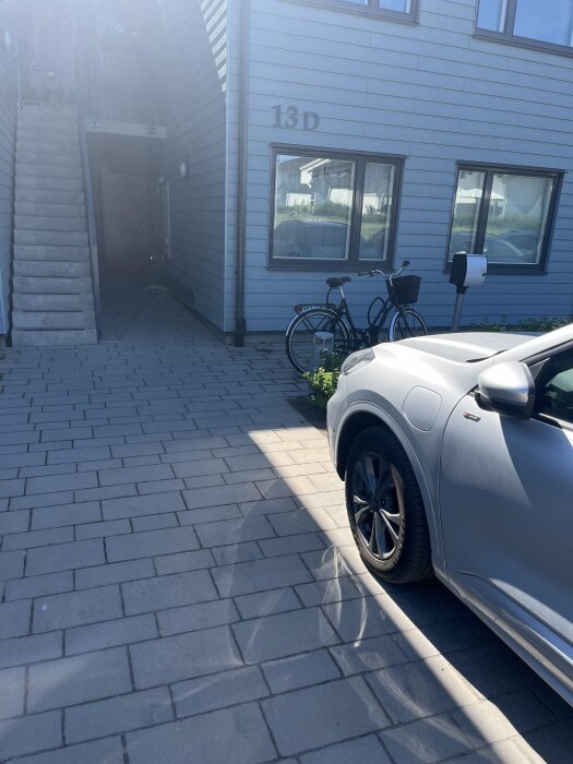 Parkeringsplats framför lägenhet, cykel parkerad vid fasad bredvid garo kupévärmare, silverfärgad bil synlig delvis från sidan.