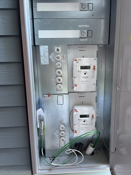 Elmätarskåp med flera strömbrytare och mätare, synlig kabeldragning och anslutningar, beläget på en husfasad.