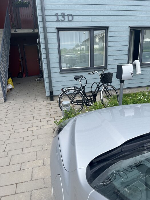 Bil parkerad framför lägenhet 13D med en cykel vid väggen och en kupévärmarstolpe bredvid.