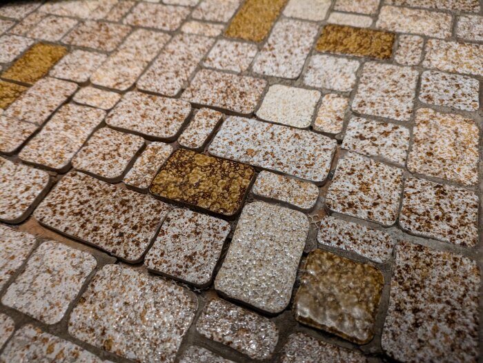 Mosaikgolv i entrén med rektangulära och kvadratiska plattor i beige och bruna nyanser, ojämnt fogade med delvis försvunnen fogmassa.