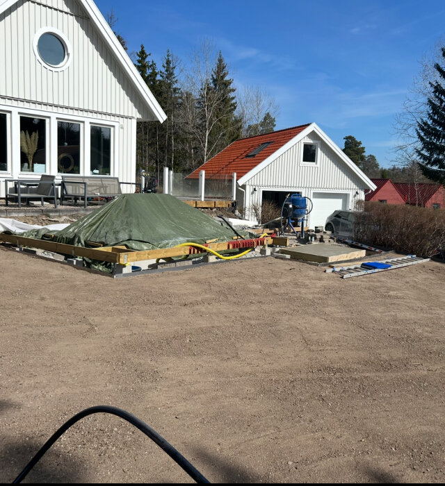 Bygge av pool i trädgård, med presenning täckt struktur, reglar och byggmaterial, vid ett hus med vitt träfasad och rött tak.
